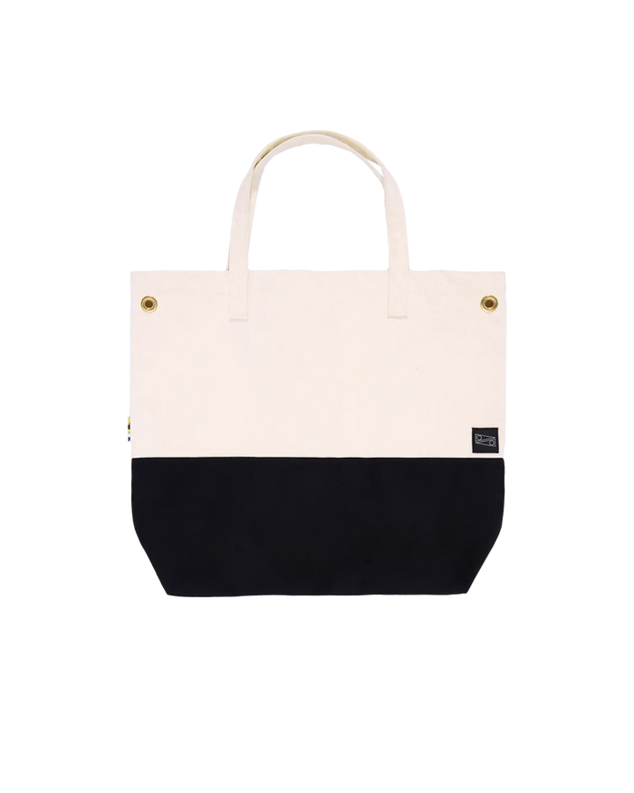 A canvas tote bag with a black bottom and white top, with brass clip closures and a small brand tag visible.
